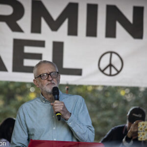 Jeremy Corbyn speaking at Palestine demo in front of stop aeming Israel banner